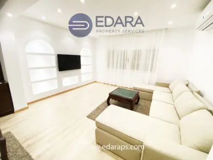 Spacious living room featuring a beige sectional sofa and a wall-mounted television with decorative shelves.