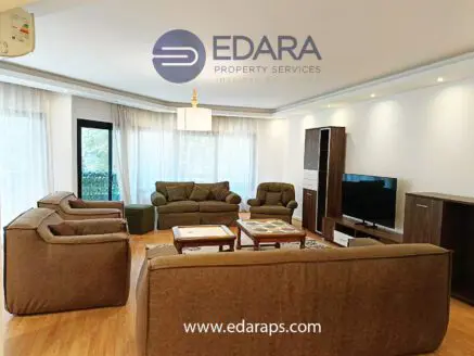Spacious living room featuring a sofa set, armchairs, and a television unit against a backdrop of large windows.