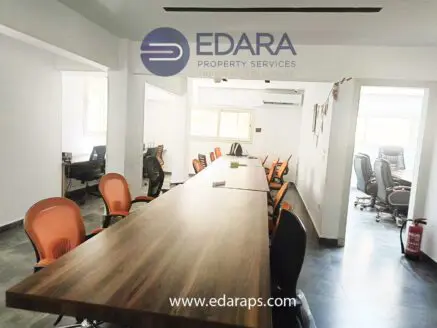 An office interior featuring a long wooden table surrounded by chairs in a well-lit space.